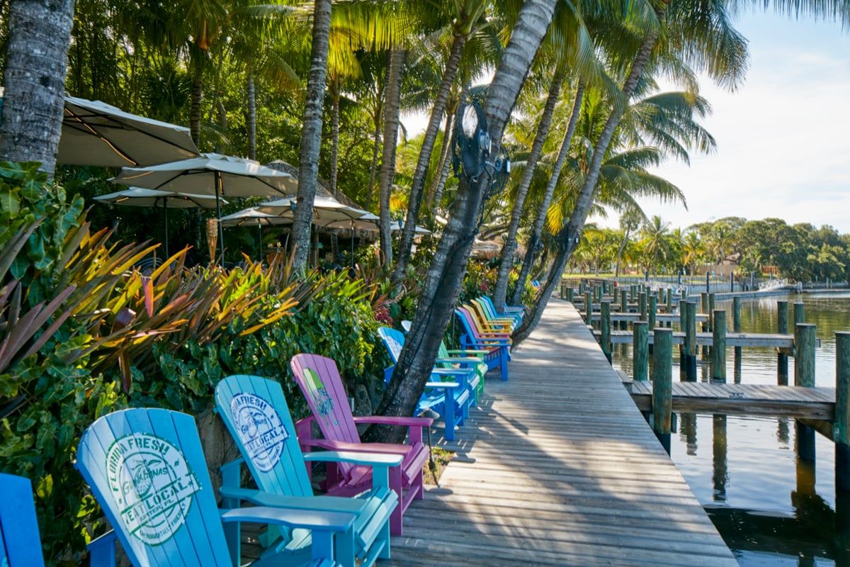 Painted Florida Fresh Adirondack chairs under leaning palms, the long wooden dock and marina pilings stretching to open water