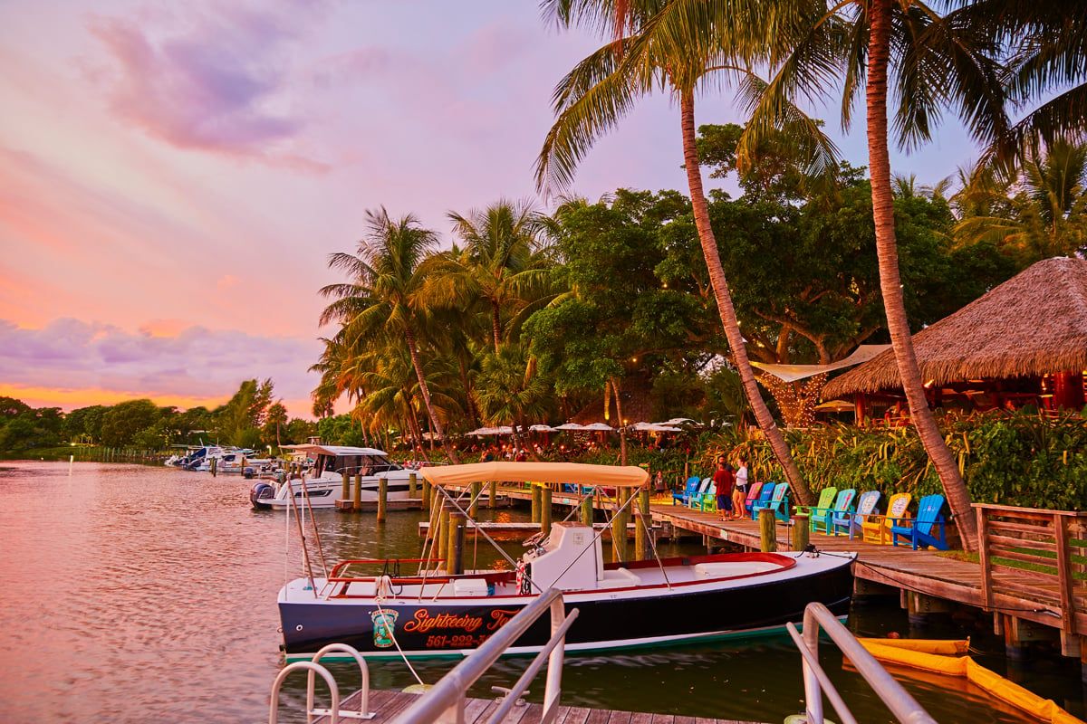 Cotton-candy pink and lavender sunset over the Intracoastal, a tour boat docked and rainbow Adirondack chairs lining the boardwalk