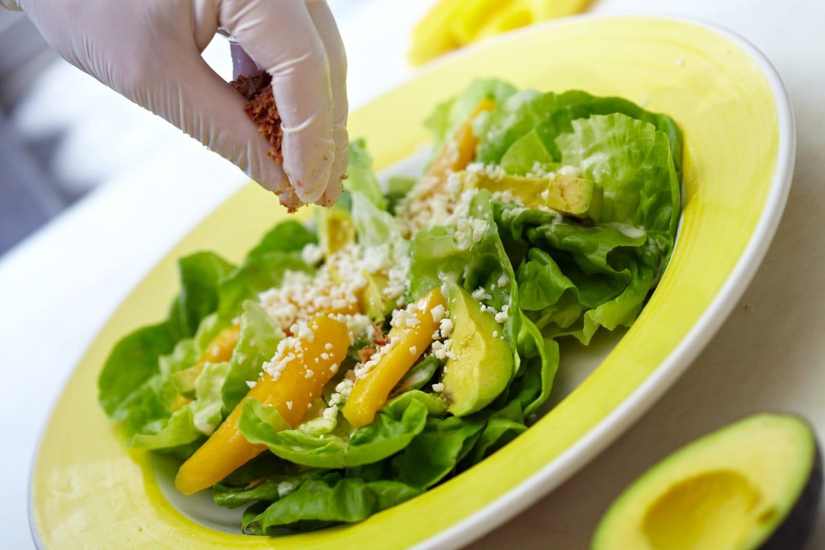 Butter-lettuce cups with mango, avocado, and crumbled cotija on a bright yellow plate, being finished by hand