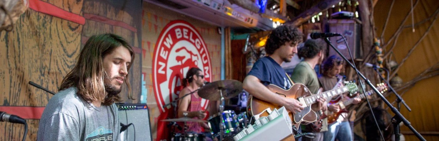 A full band jamming on the indoor tiki stage at night — keys, drums, and two guitarists under warm light with a real crowd